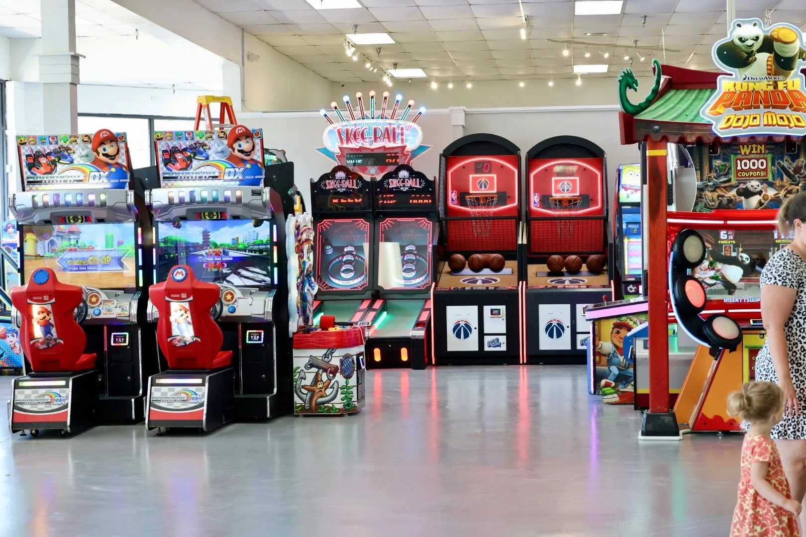 A row of arcade games in an indoor arcade, including Mario Kart with red seats, basketball shooting games, and a Kung Fu Panda game, with a woman and a small child standing nearby.