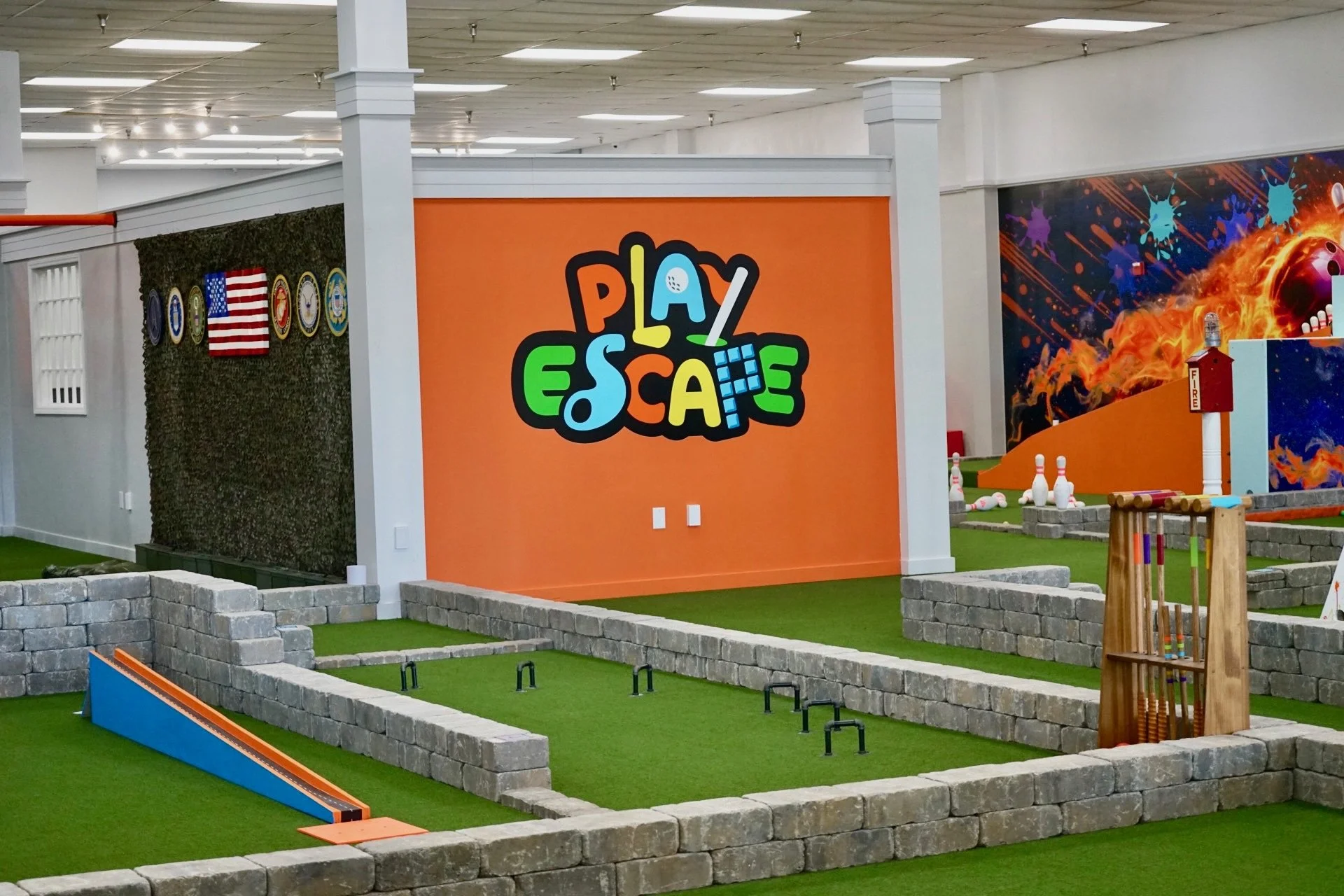 Indoor mini golf area with artificial green turf, small stone walls, and a large orange sign reading 'Play Escape'. The background features a wall with a space-themed mural and protest flags of the United States, Marine Corps, Navy, and Army.