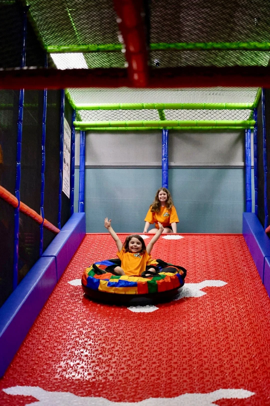 Two young girls wearing orange shirts playing on a red, padded indoor playground slide with white paw prints, enclosed by multicolored padded walls and climbing netting.
