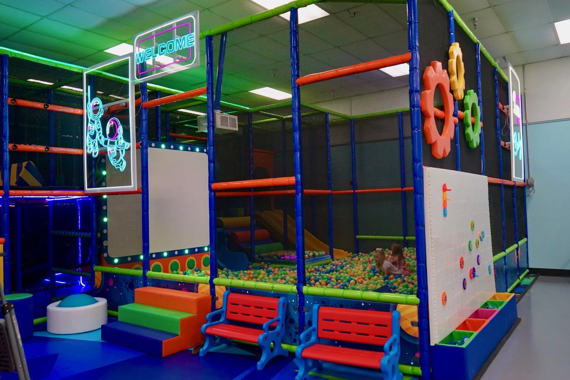 Indoor children’s play structure with climbing areas, slides, and ball pit, decorated with colorful gears and neon signs that say 'Welcome' and display an astronaut illustration.