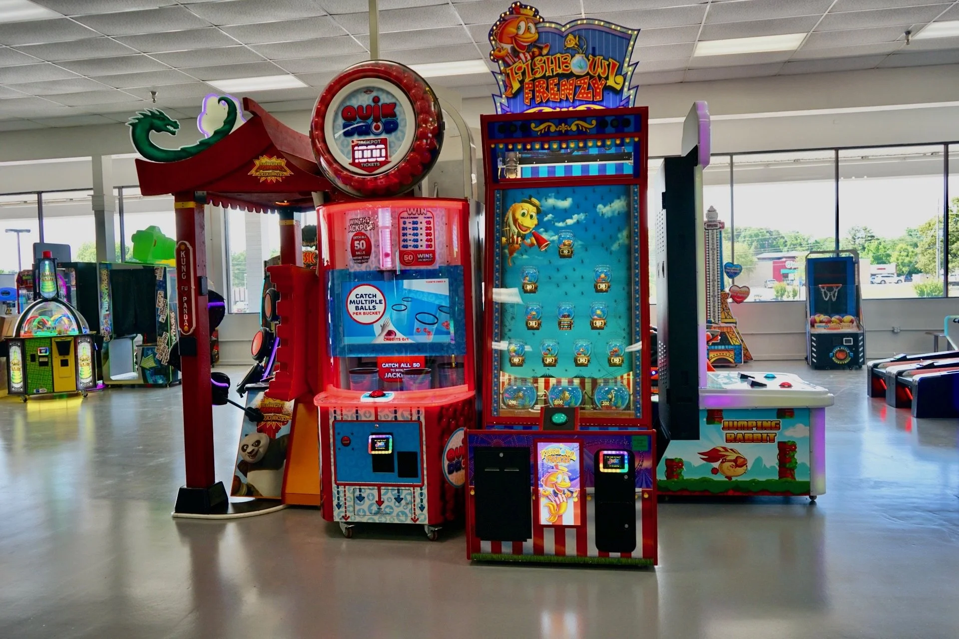 Indoor arcade with various claw and ticket redemption games, including a Frog Jumping Rabbit game, a shooting fish frenzy game, and a gumball machine, in a brightly lit space with windows in the background.