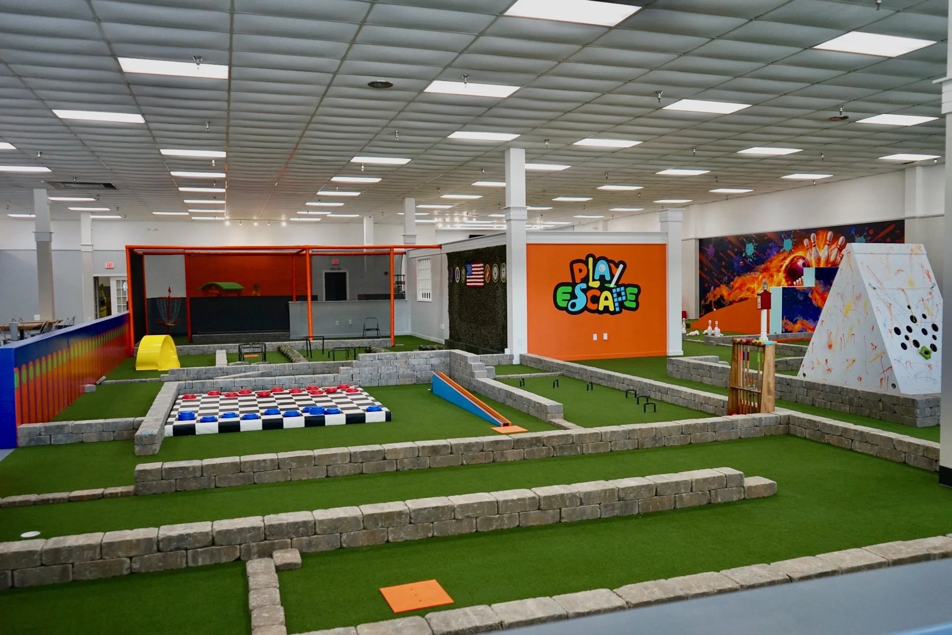Indoor mini golf course with artificial grass, stone borders, and various game obstacles. Features a colorful sign reading 'Play Escape,' an orange wall, and a large vibrant mural in the background