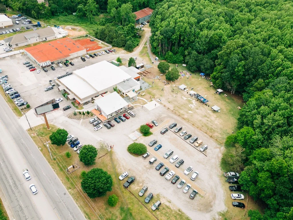 An aerial view of a commercial building with parking lots, surrounded by trees and a grassy area, with a road on the left side.