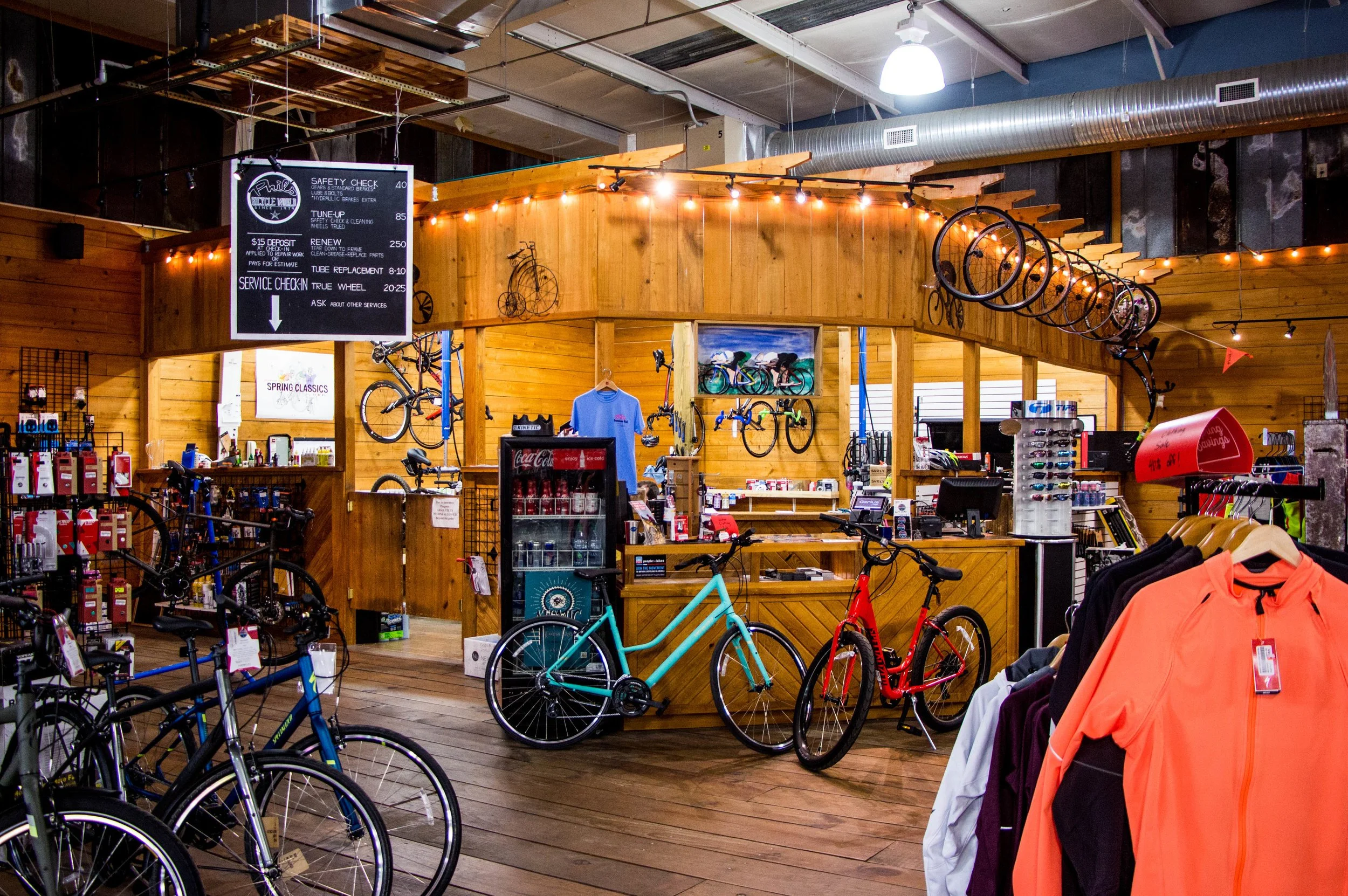 Interior of a bike shop with bicycles on display, wooden walls, and a service counter. Bicycles and cycling apparel are visible, along with a digital signboard and a Coca-Cola fridge.