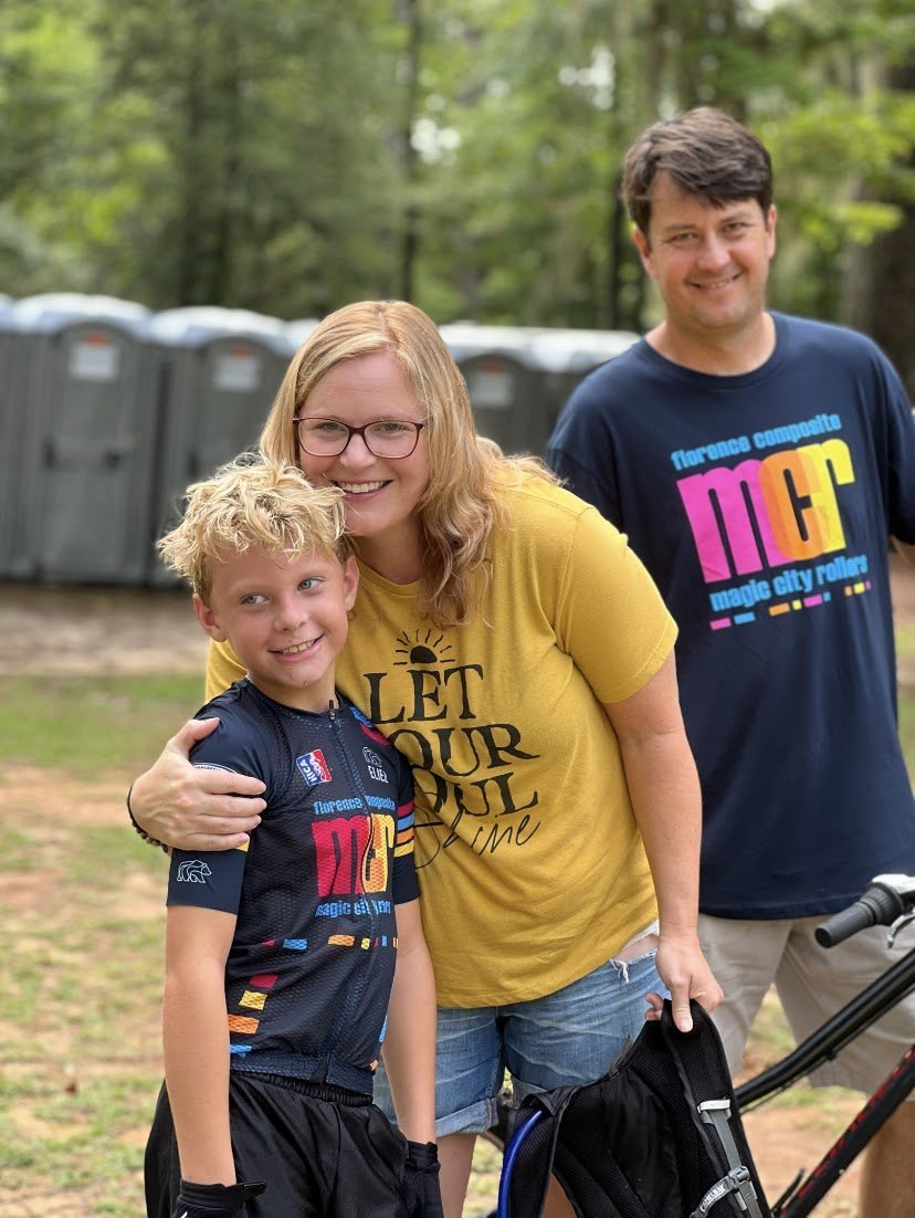 Three people outdoors, a woman and two boys, smiling at the camera, with portable toilets in the background.