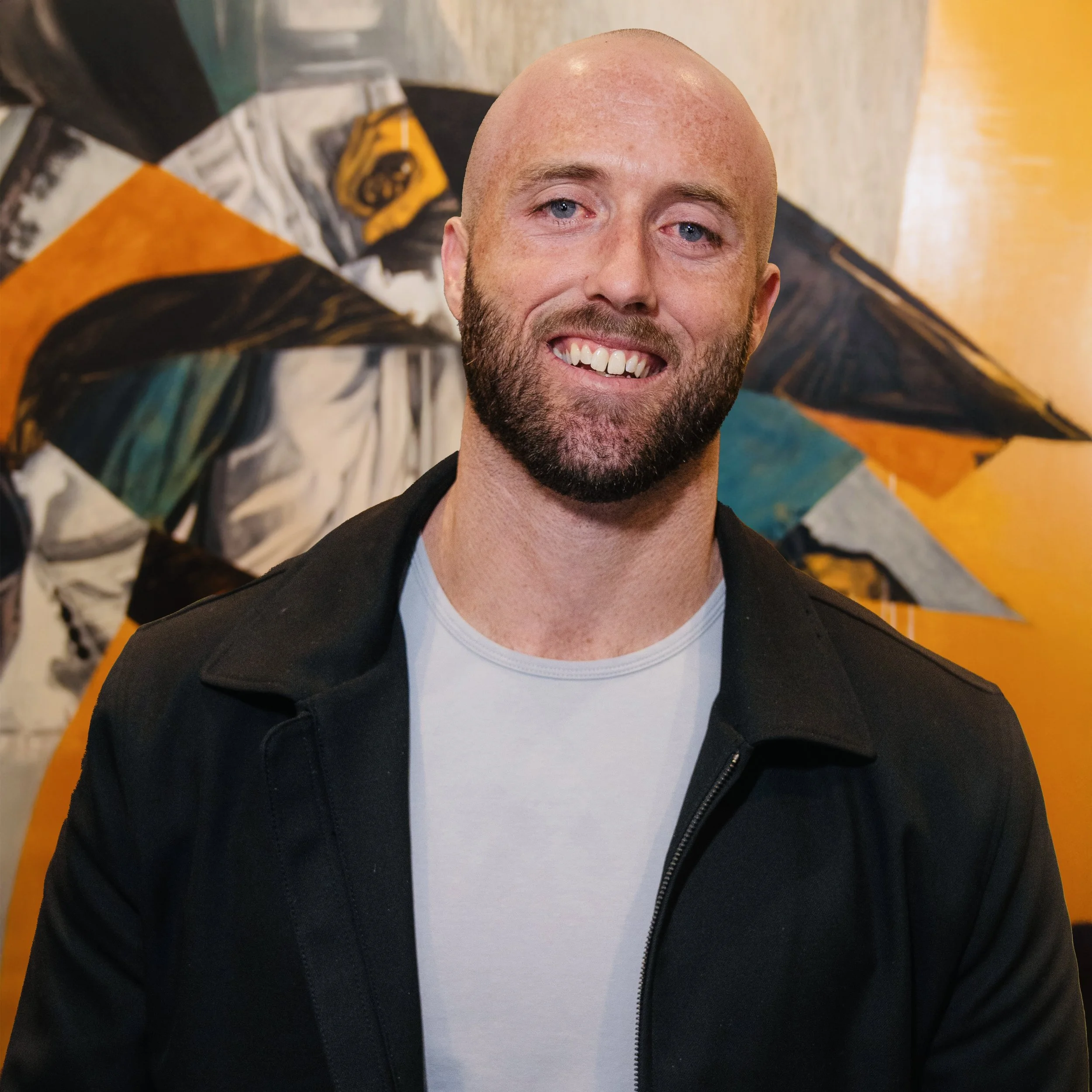Portrait of a smiling man with a beard and bald head, wearing a black jacket over a white t-shirt, standing in front of colorful abstract art.