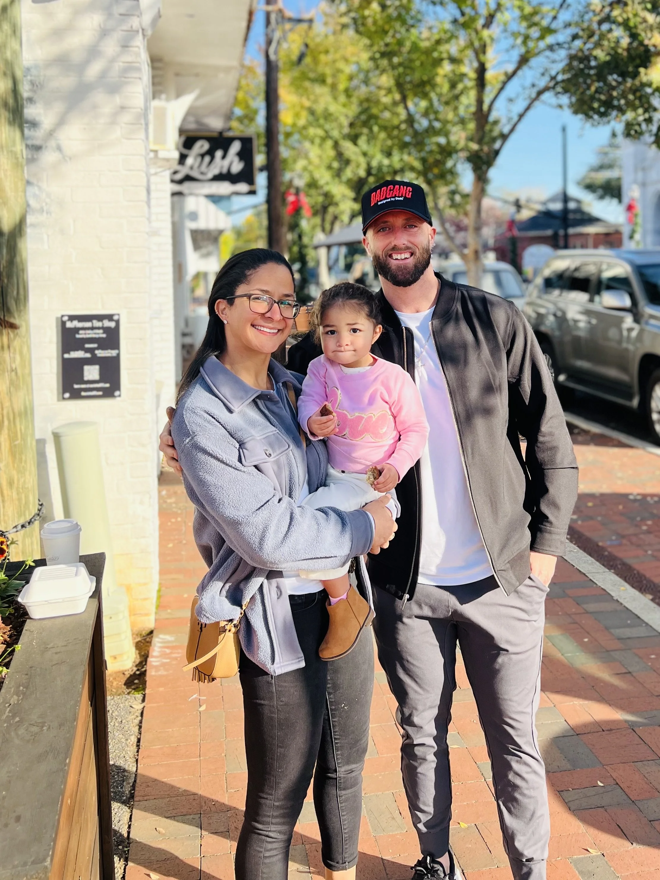 A happy family of three, including a woman, a young girl, and a man, smiling and standing outdoors on a sunny day near a brick sidewalk, trees, parked cars, and a storefront with a sign that reads 'Lush' in the background.