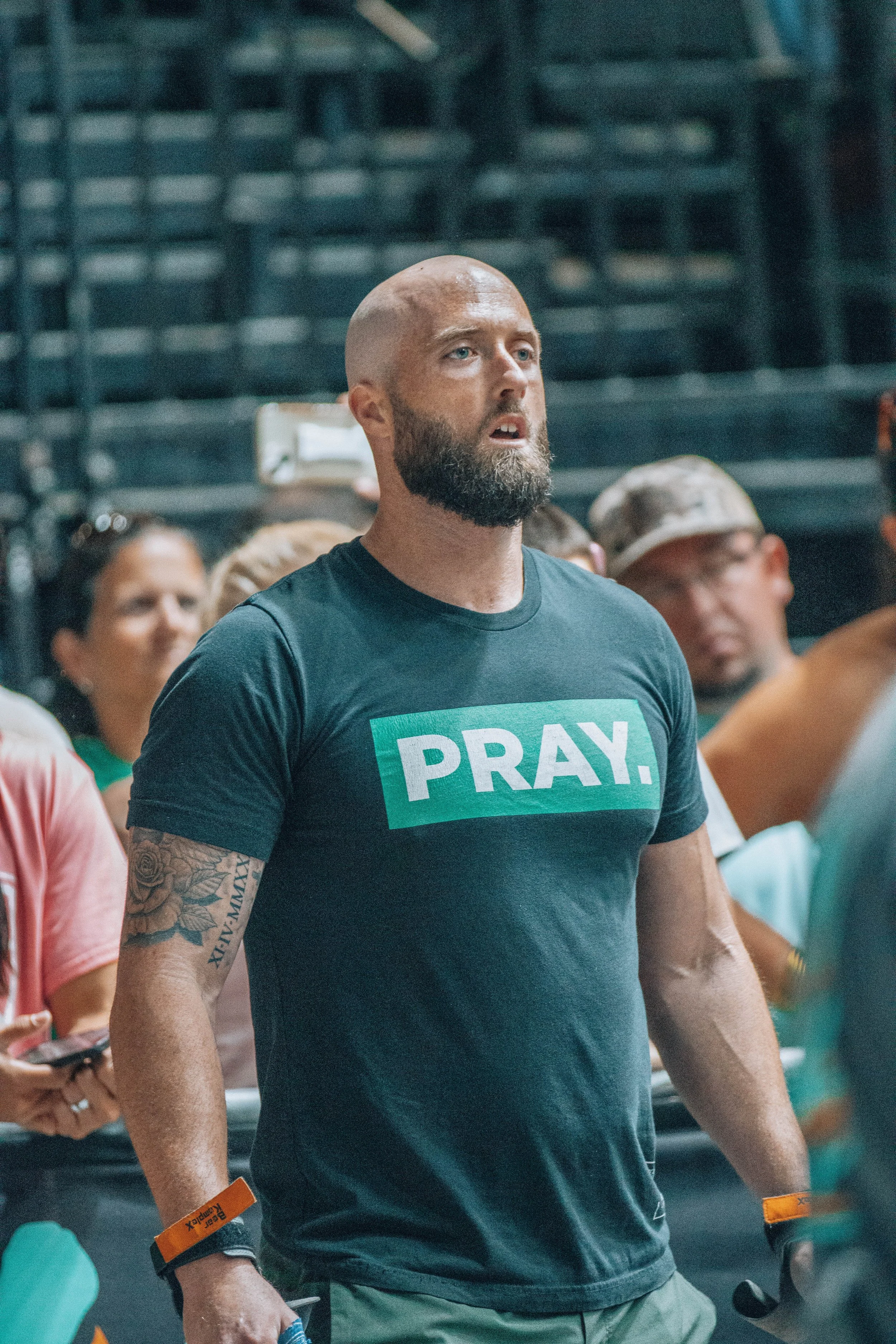 A man with a shaved head and beard wearing a black T-shirt with the word "PRAY." on it, standing among a crowd.