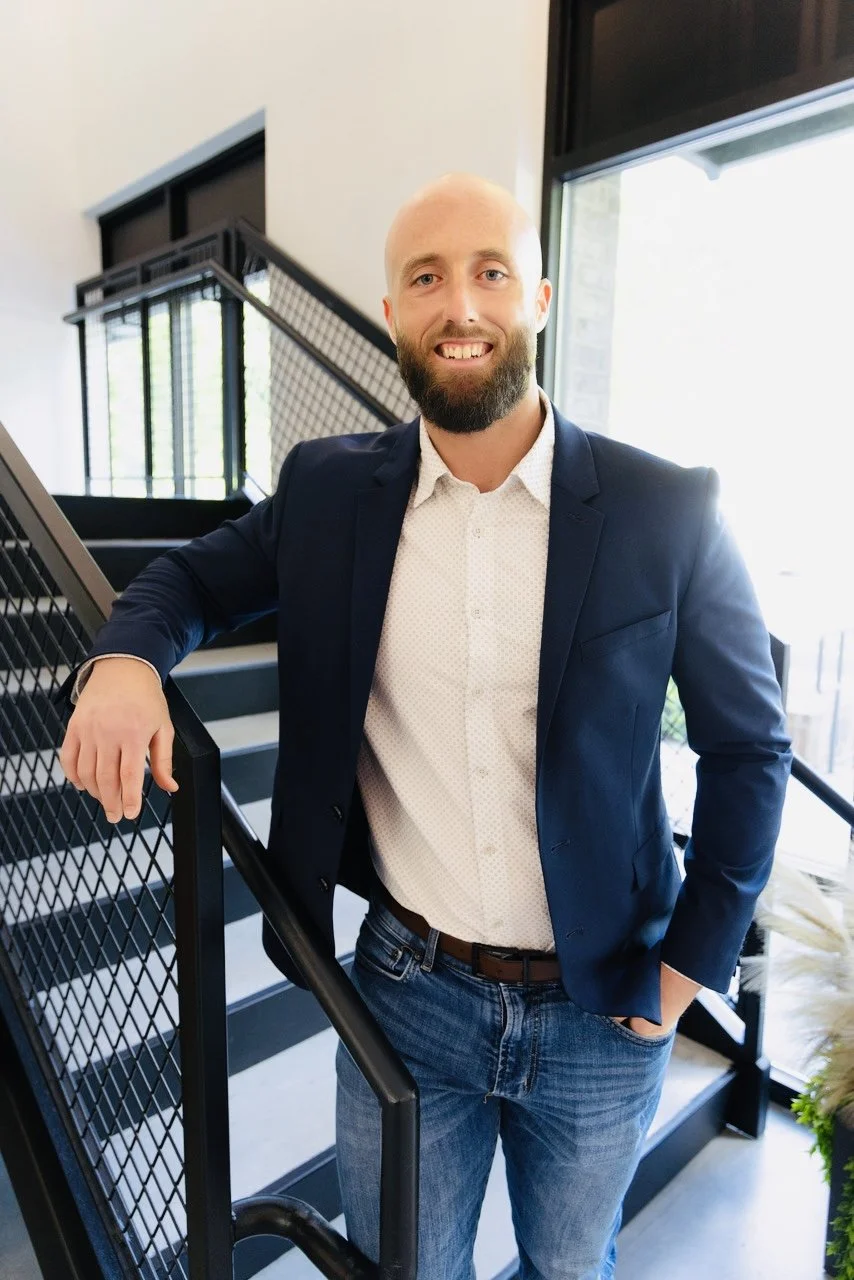 Man with a beard and glasses smiling, standing on a staircase indoors, wearing a navy blazer, white shirt, and jeans.