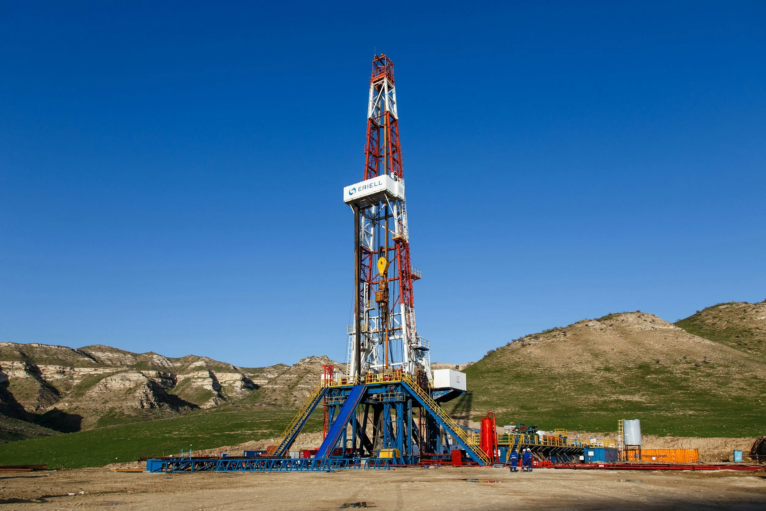 An oil drilling rig located in a mountainous area with green hills and a clear blue sky.