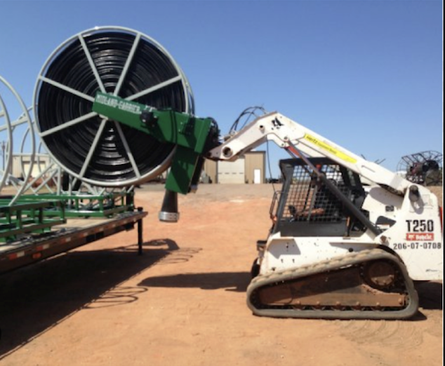 A white and black tracked construction robot with a hydraulic arm is lifting a large green irrigation hose reel on a flatbed trailer in an outdoor yard during daytime.