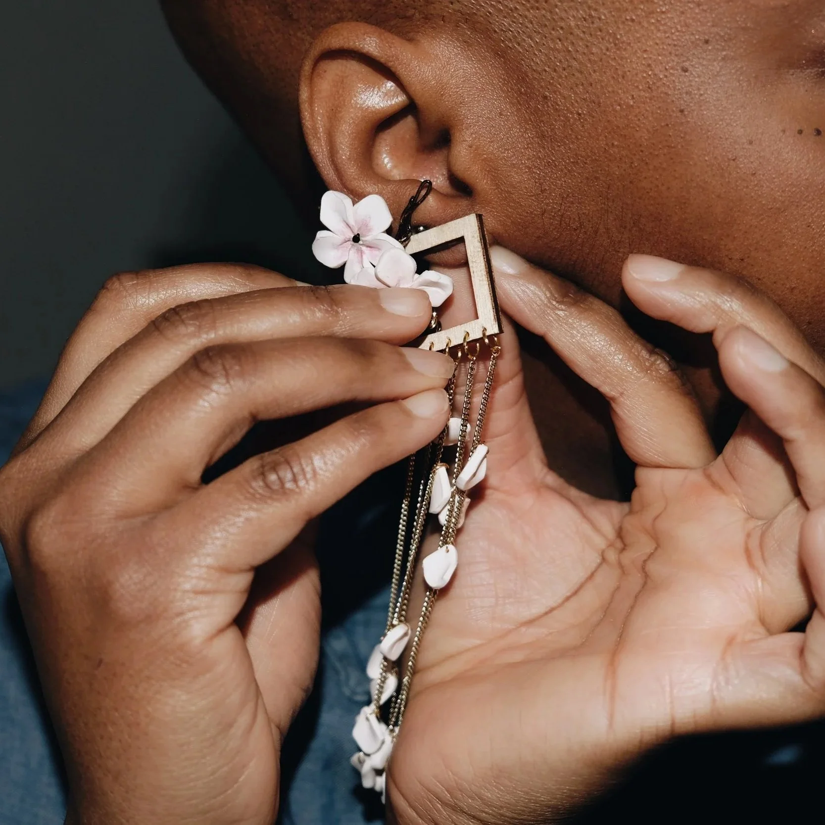 Close-up of a person with dark skin holding an intricate earring with pink flowers and white shell details near their ear.