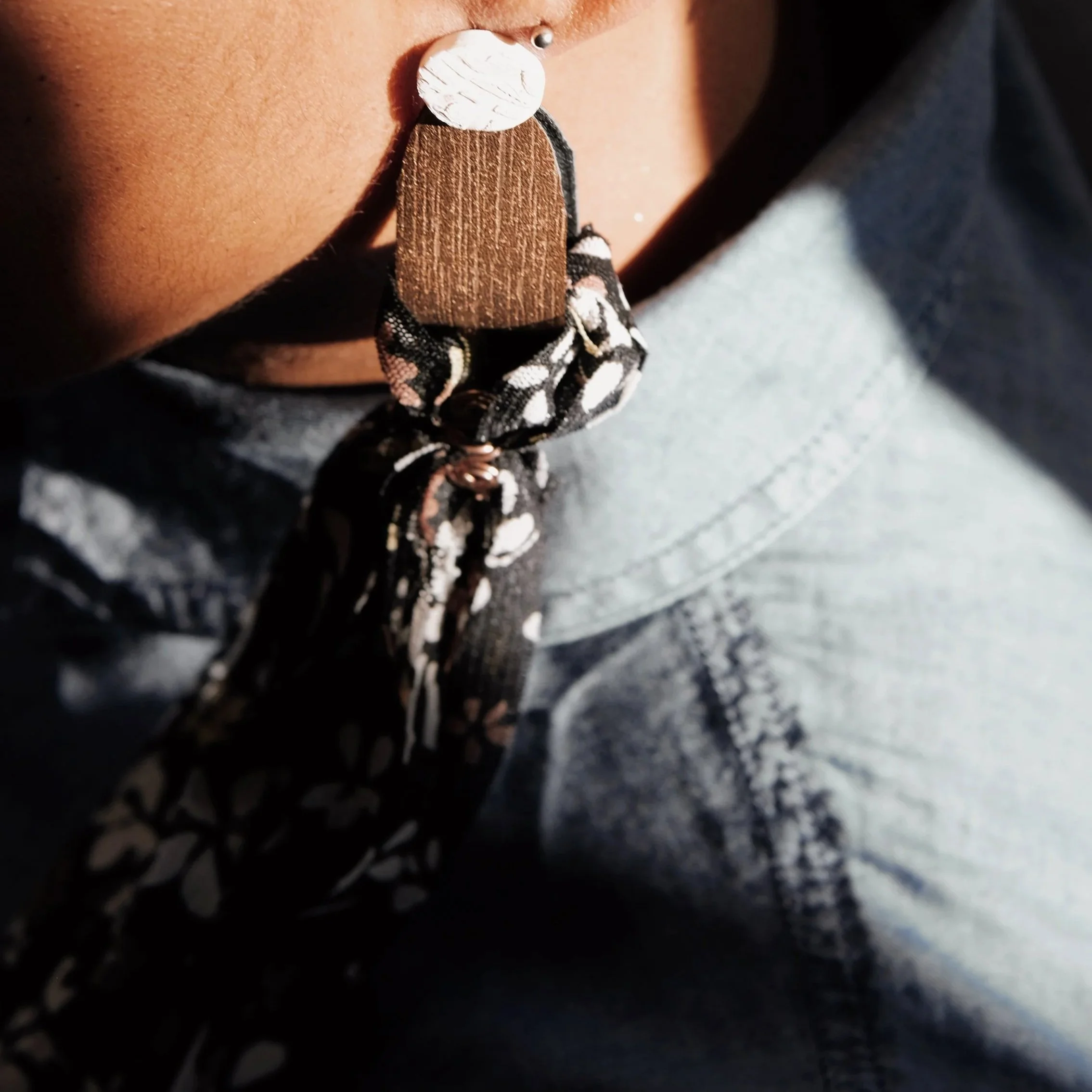 Close-up of a person's neck and chest wearing a necklace with a large bead, wooden pendant, and black-and-white patterned scarf, with a gray top.