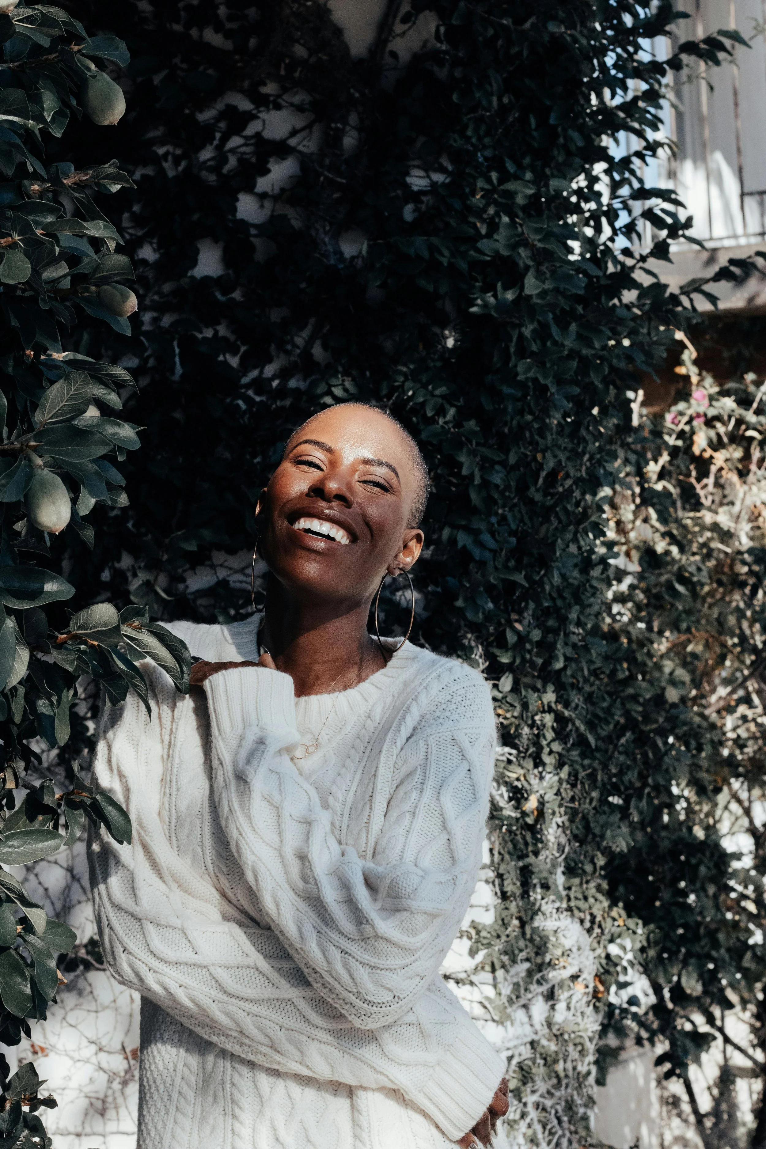 A smiling woman in a white cable-knit sweater standing outdoors among green and snow-covered foliage.