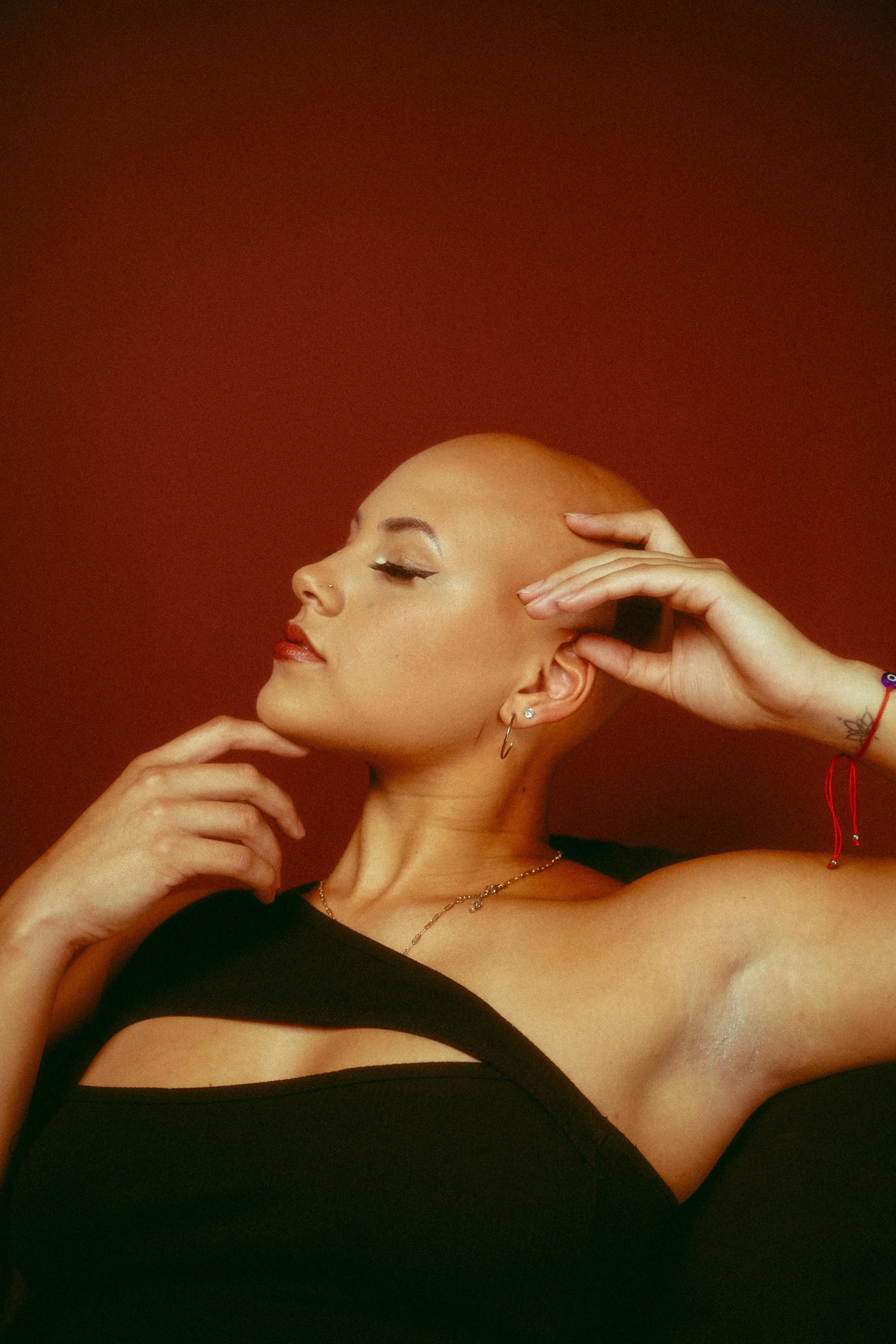 A woman with a shaved head poses with her eyes closed, wearing makeup, earrings, and a black top with an open shoulder design, against a red background.