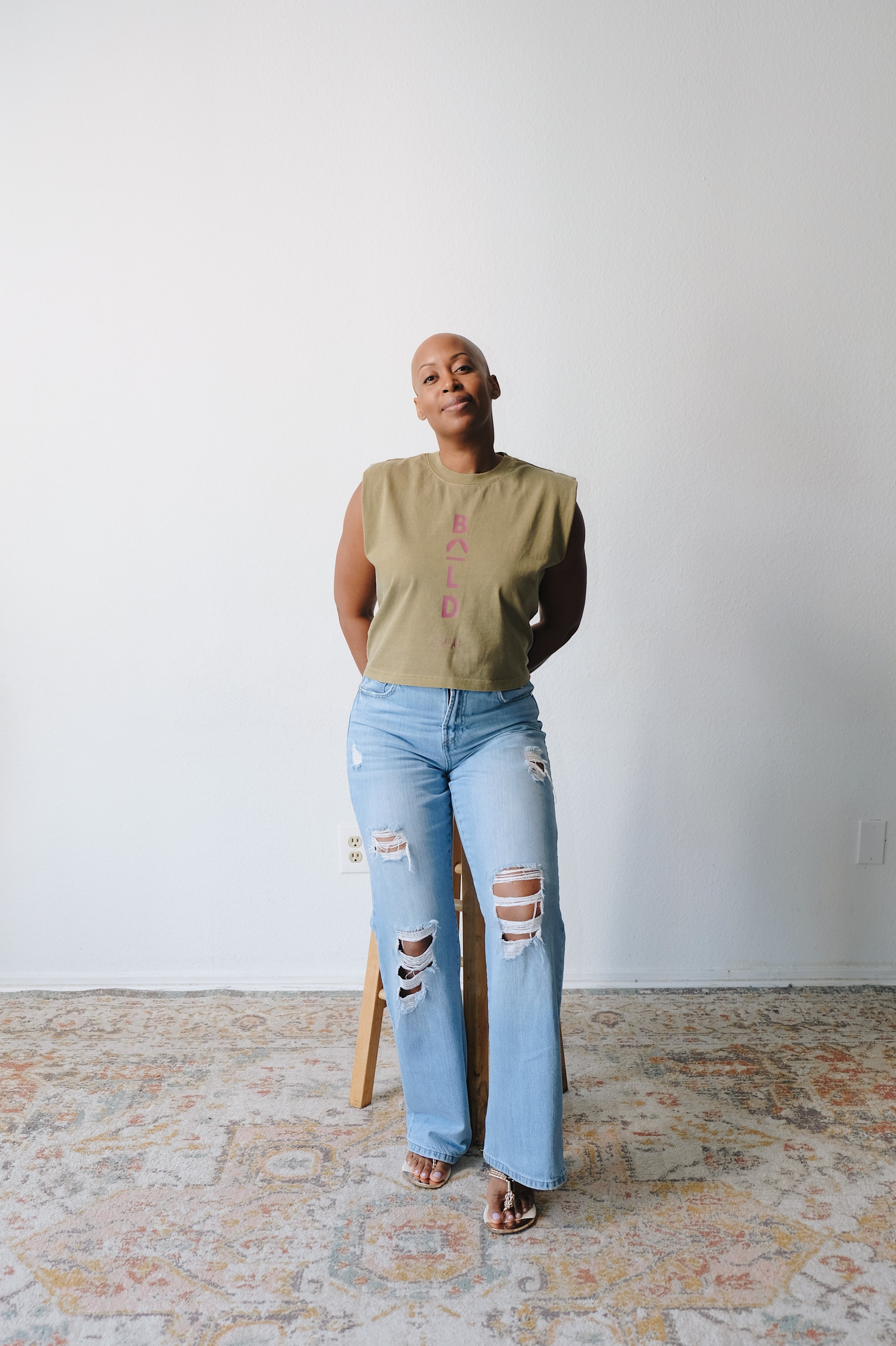 A woman standing against a plain white wall, wearing a sleeveless olive green shirt with the word 'BUILD' written vertically in pink, ripped jeans, and open-toed heels.
