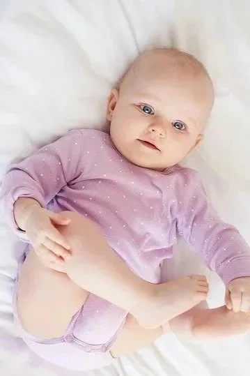 Baby lying on a white bed, wearing a pink long-sleeve shirt with small white dots, looking at the camera.