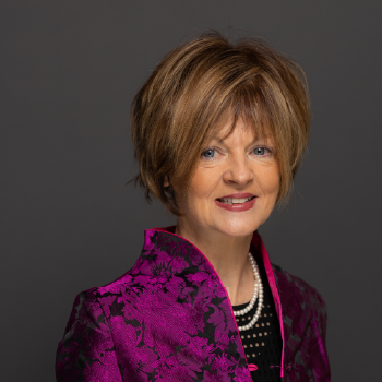 Headshot of a middle-aged woman with short, layered brown hair, wearing a purple patterned blazer and pearl necklace against a gray background.