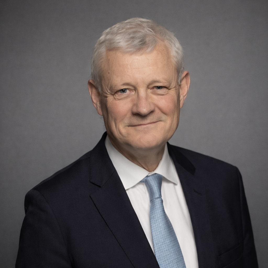 Portrait of an older man wearing a dark suit, white shirt, and light blue tie, against a gray background.