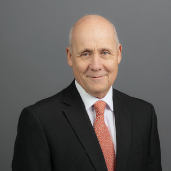 An older man in a black suit, white shirt, and orange tie, smiling against a gray background.