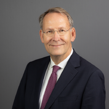 Portrait of a middle-aged man in a suit and tie, smiling against a gray background.