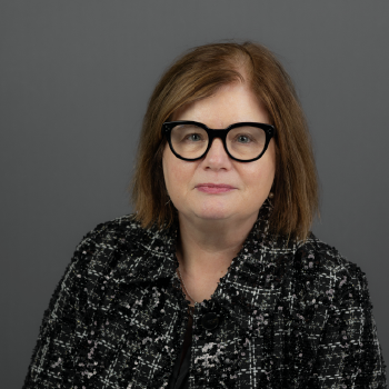 A woman with shoulder-length red hair and black glasses, wearing a black and white patterned blazer, posing in front of a plain gray background.