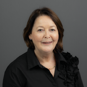 A middle-aged woman with dark brown hair styled in a bob, wearing a black blouse with ruffle details, smiling against a gray background.