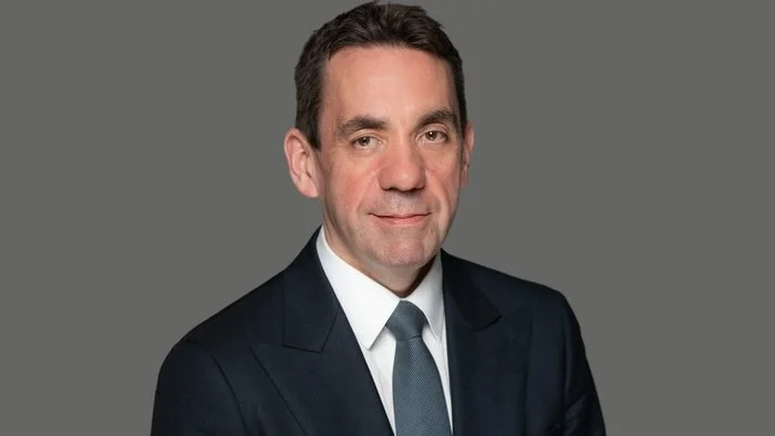 Professional headshot of a middle-aged man in a dark suit, white shirt, and tie, with a neutral gray background.