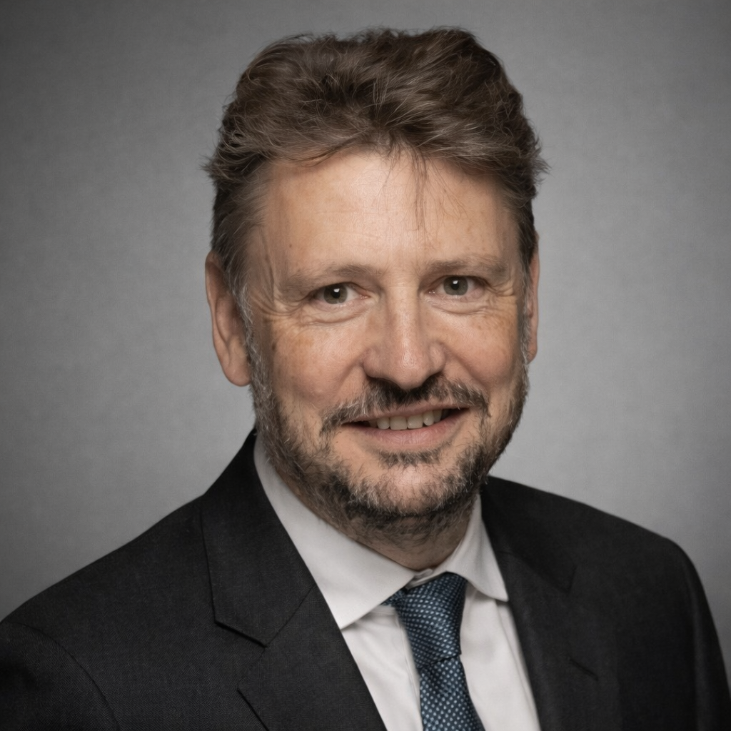 Headshot of a middle-aged man with light brown hair, beard, wearing a black suit, white shirt, and blue tie, smiling against a gray background.