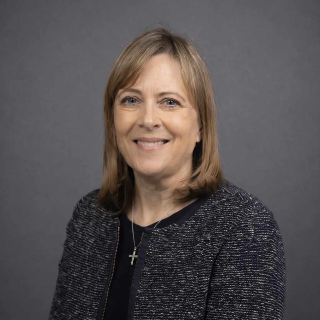 Portrait of a smiling woman with shoulder-length light brown hair, wearing a dark patterned blazer and cross necklace, against a gray background.