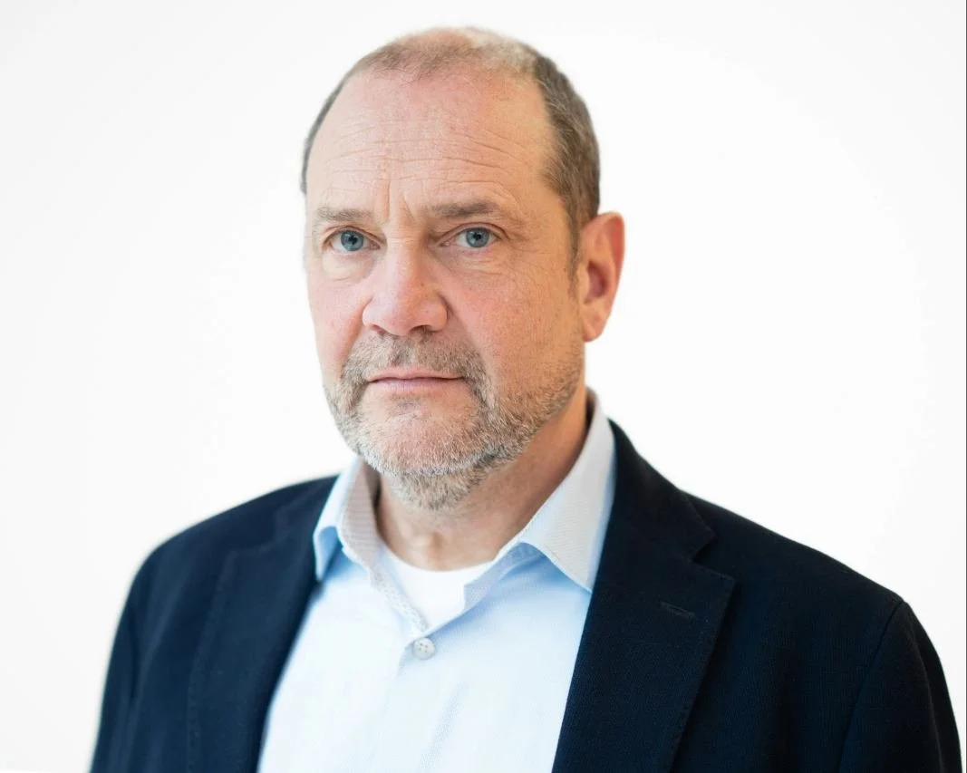 A middle-aged man with short gray hair and blue eyes, wearing a dark blazer and a white shirt, against a plain white background.