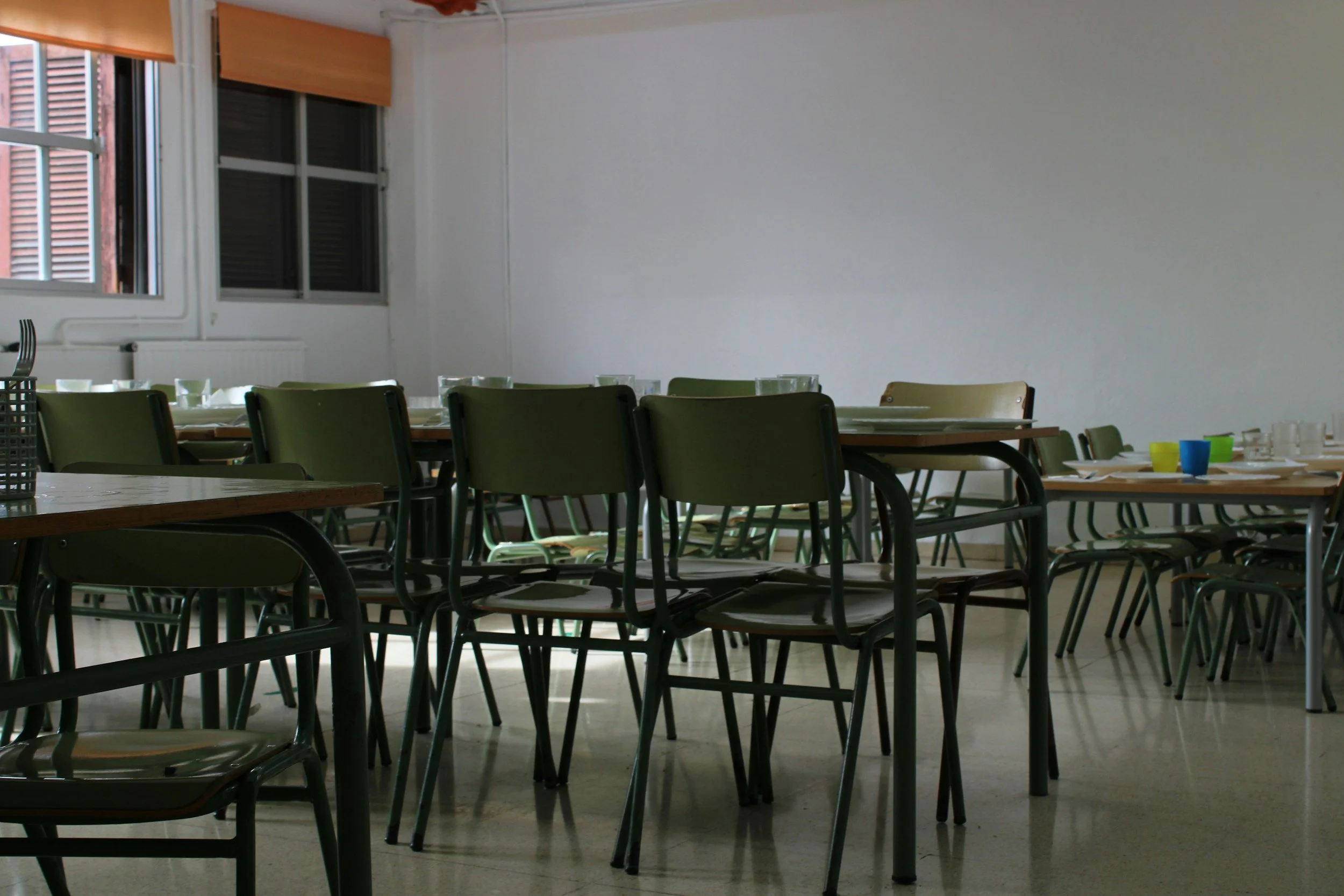 Empty dining room with tables and chairs, set with cups and plates, near windows with blinds.