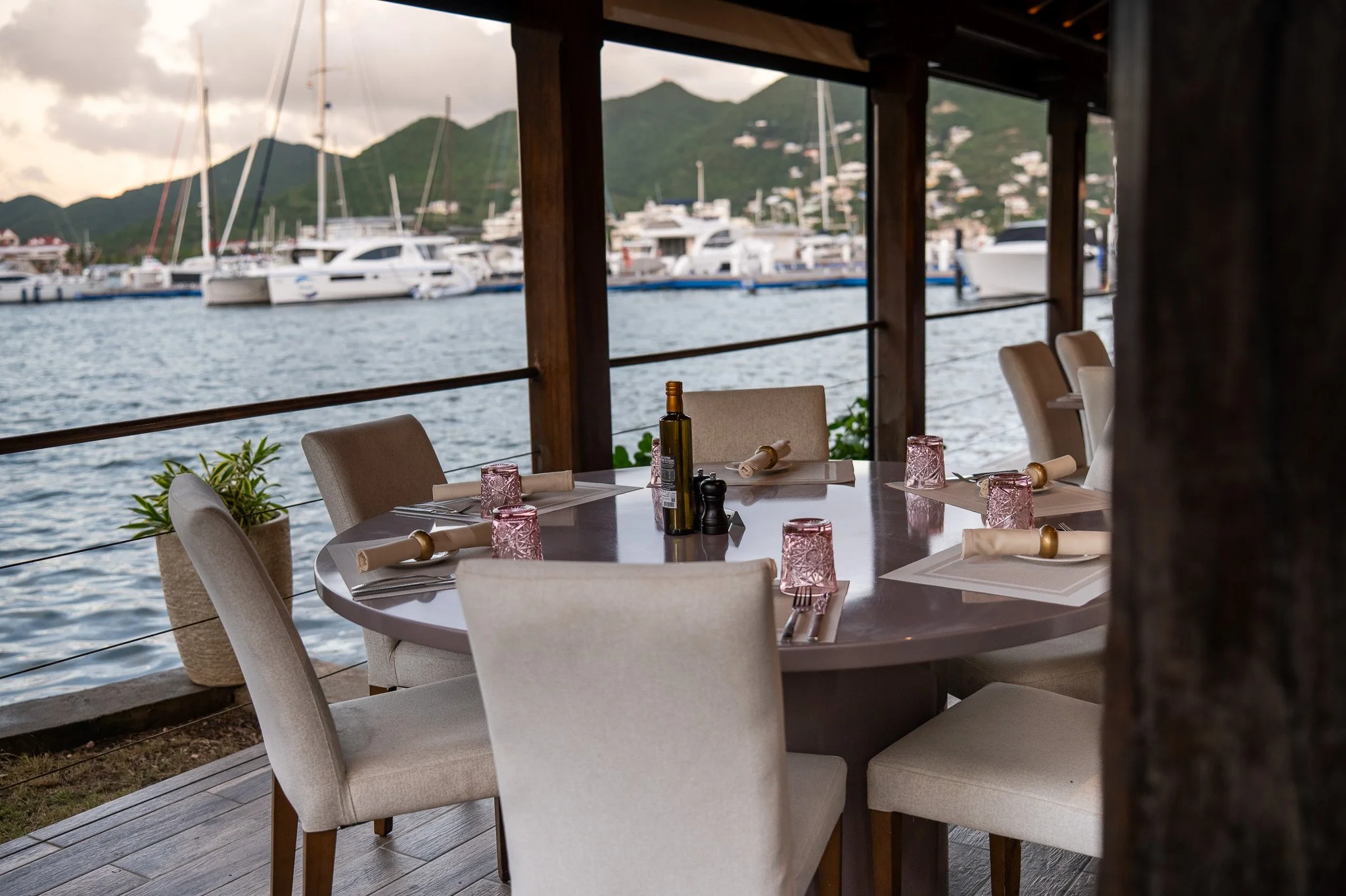 Table set for dining with placemats, napkins, and glasses, overlooking a marina with yachts and green mountains in the background.