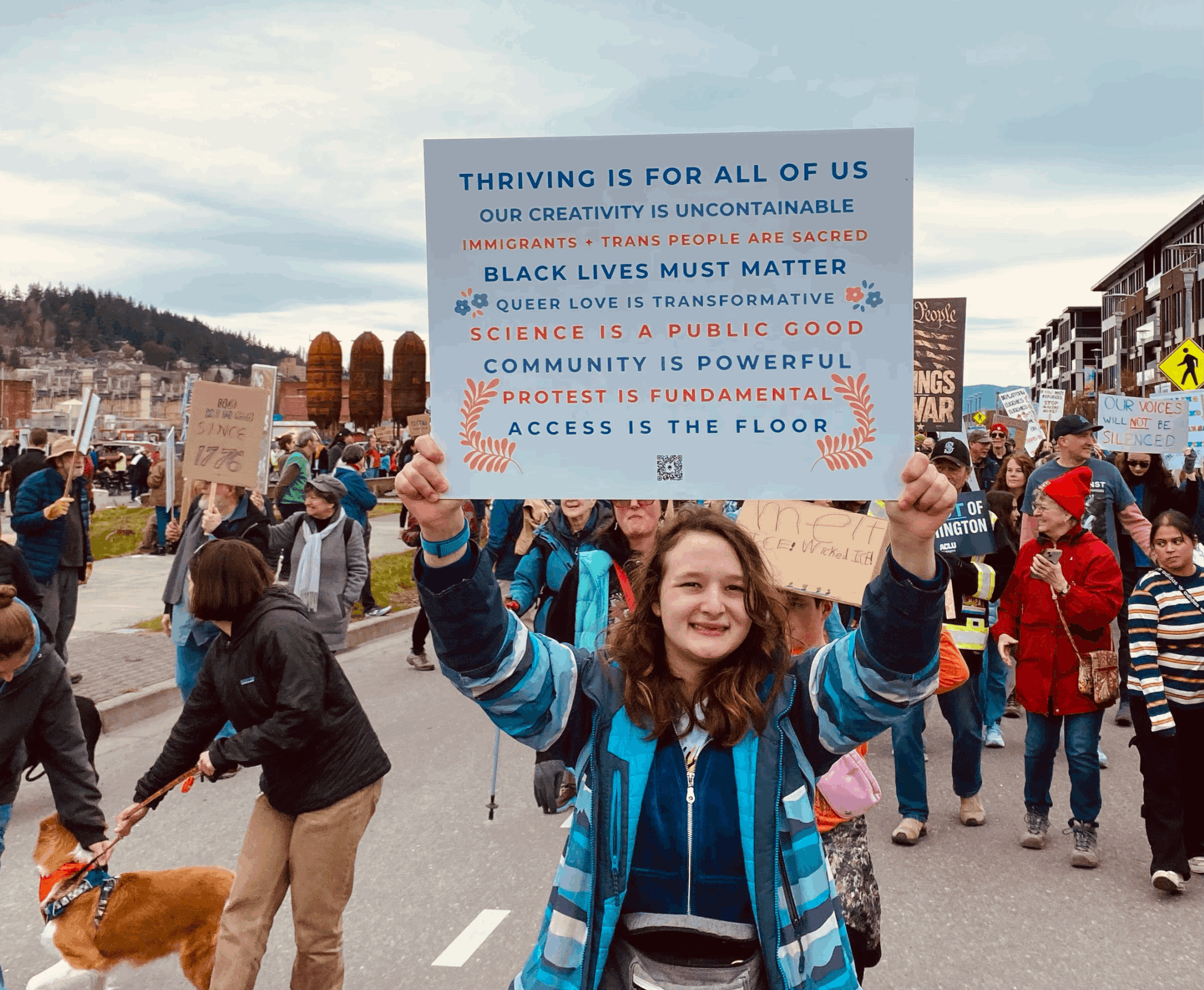 Photo of a young person holding Thriving Is For All Of Us yard sign at a protest
