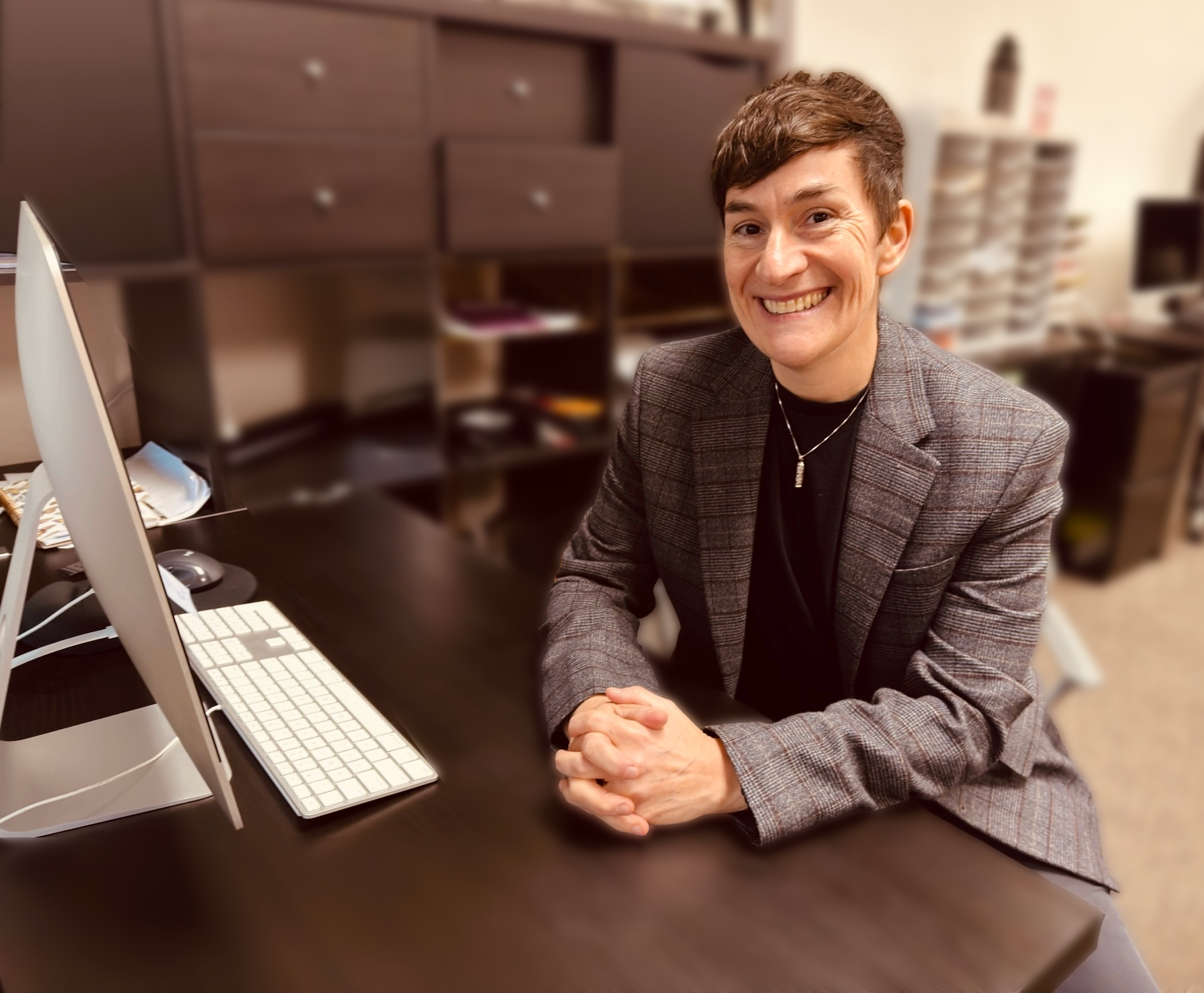 Litav is sitting at a dark wood desk with a computer monitor and keyboard. They are smiling warmly and wearing a dark colored plaid blazer.
