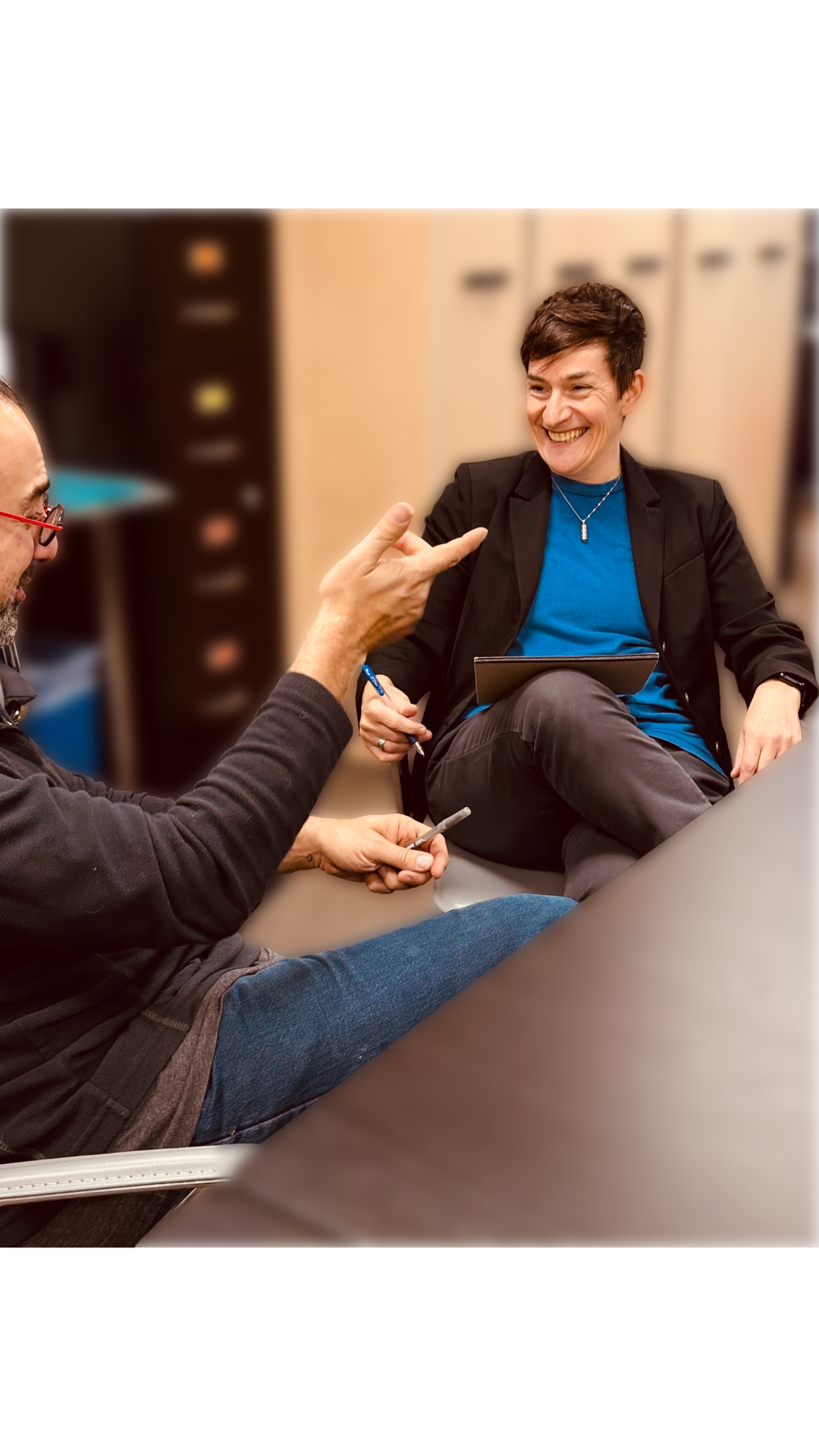 Litav and another person sit at an office table having a conversation, smiling. The person Litav is talking with is gesturing happily.