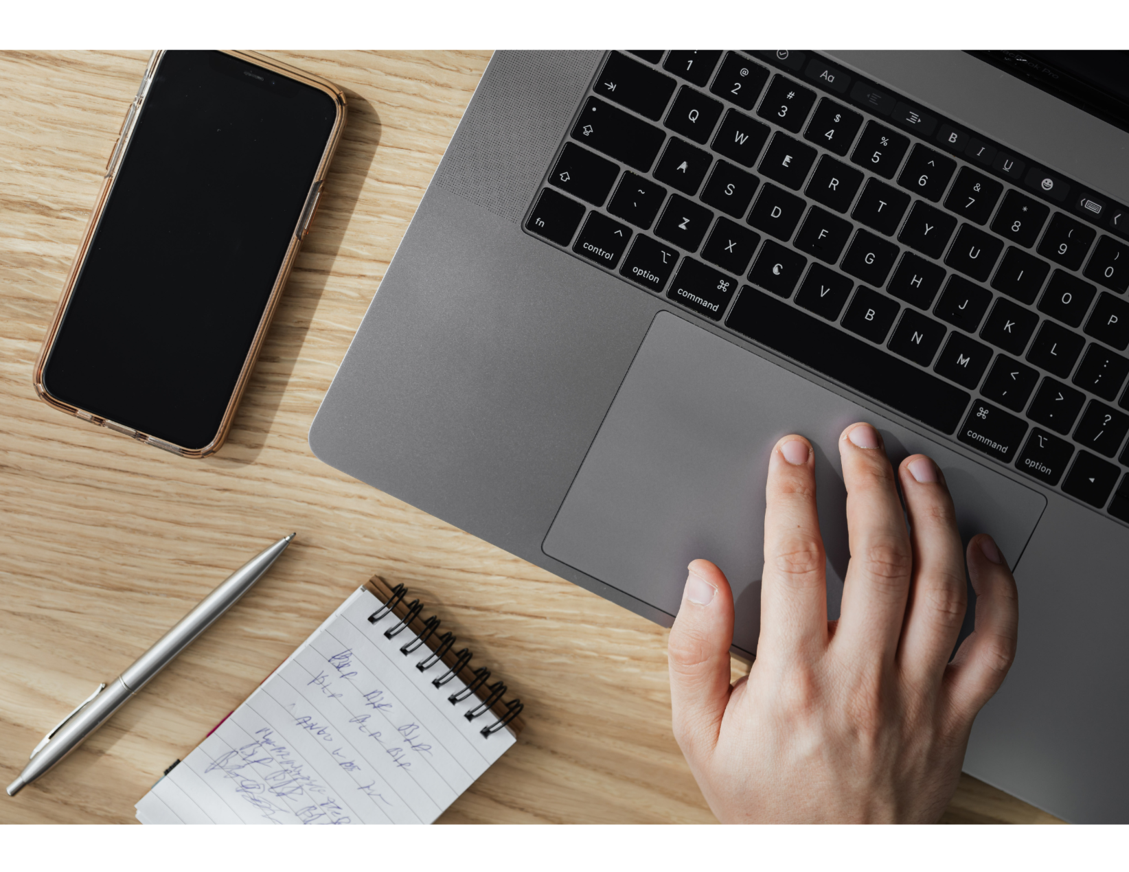 Photo of laptop keyboard with hand on mousepad, plus phone, small pad of paper, and pen.
