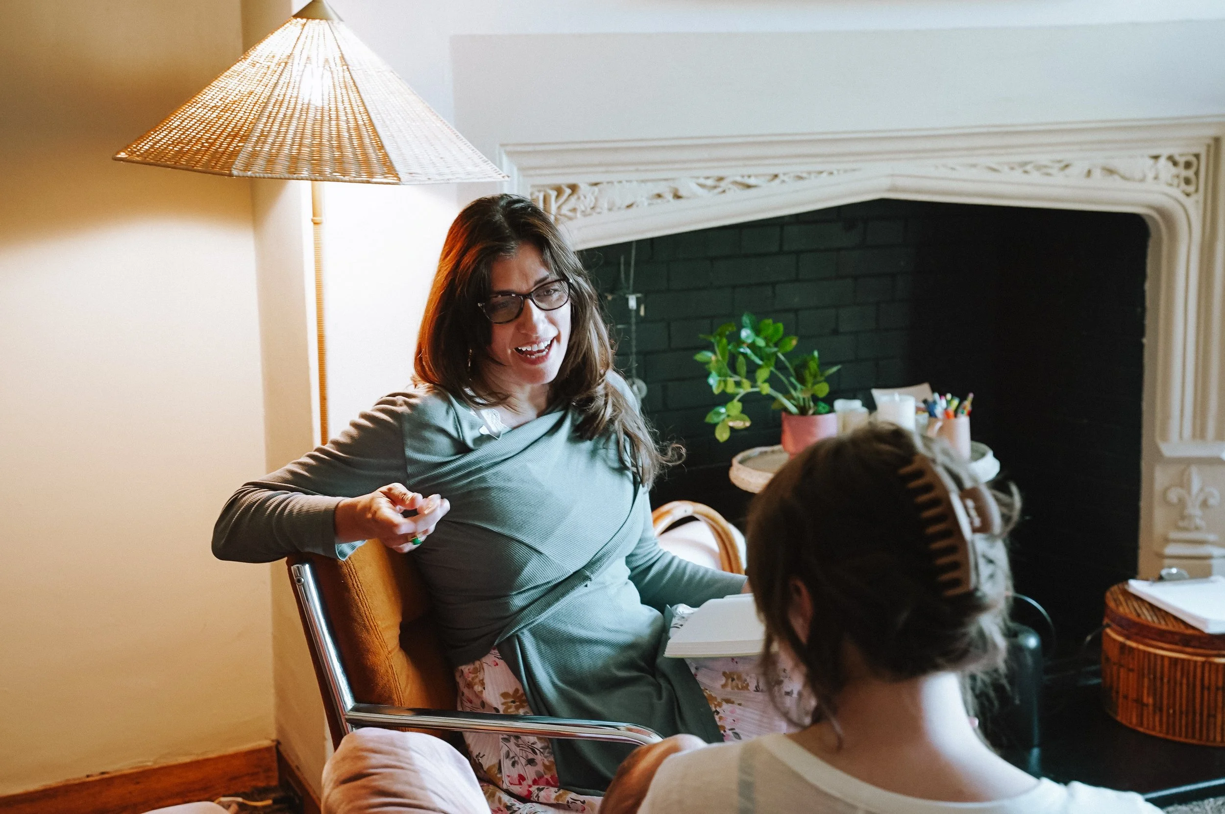 A woman with long hair and glasses smiling while sitting at a table during a gathering, with children nearby, in a room with a fireplace and decorative molding.