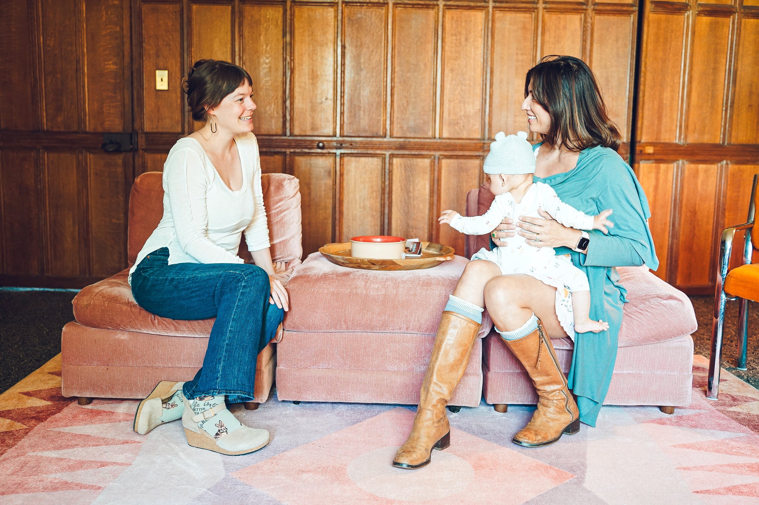 Two women and a baby sitting on a pink couch in a wood-paneled room, smiling and talking to each other.