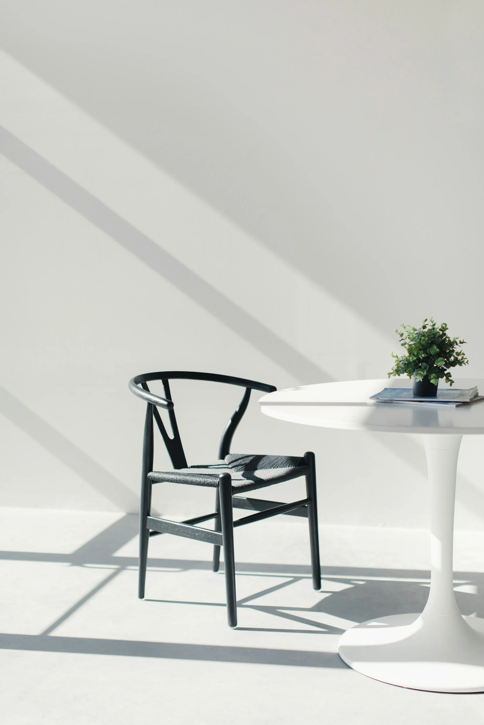 A modern black chair and a white round table with a small potted plant and stacked magazines, all illuminated by sunlight with shadows on a white wall.