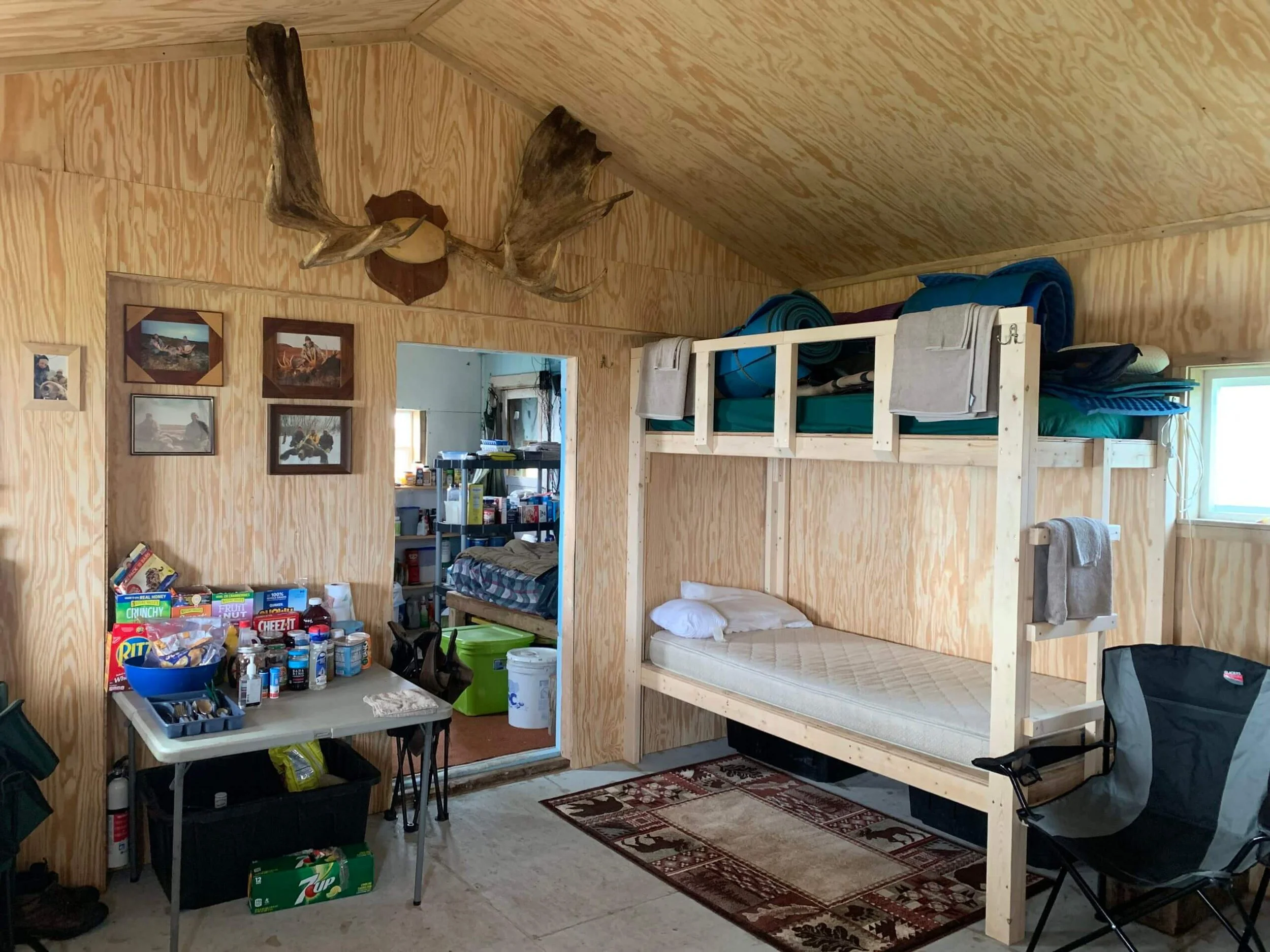 Interior of a rustic cabin room with wooden walls and ceiling, featuring a moose antler mounted on the wall, photos and artwork, a workplace table with supplies, a bunk bed with camping gear, and a camping chair next to a window.