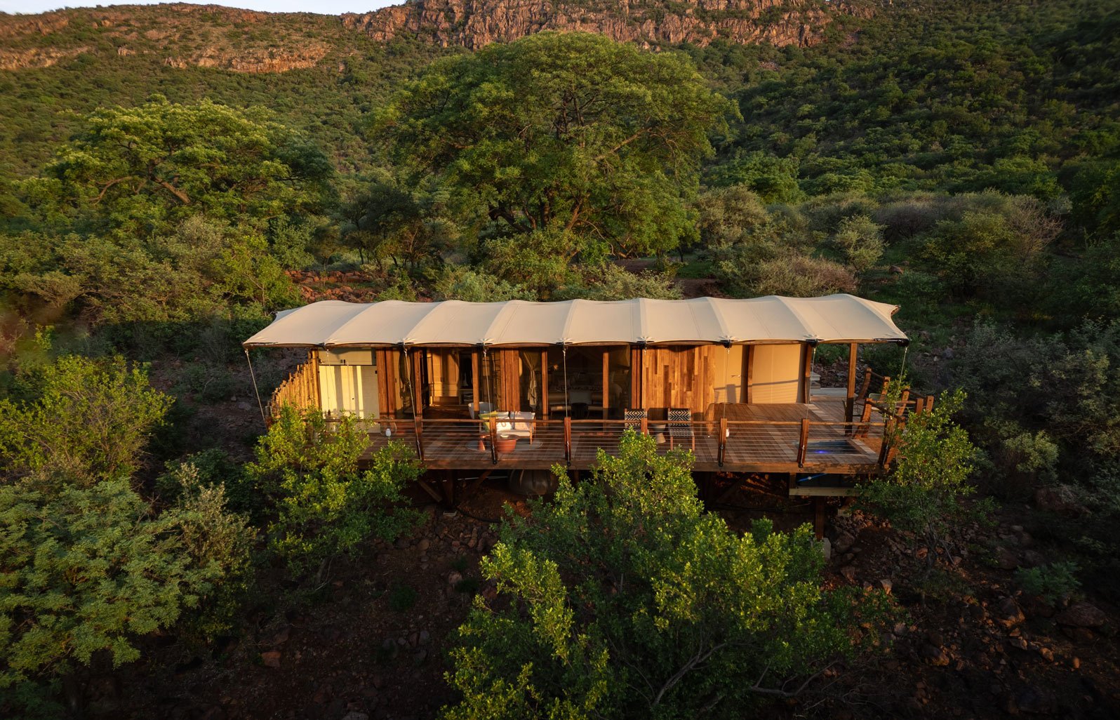 A raised wooden cabin with a fabric roof is situated among green trees in a mountainous area at sunset.