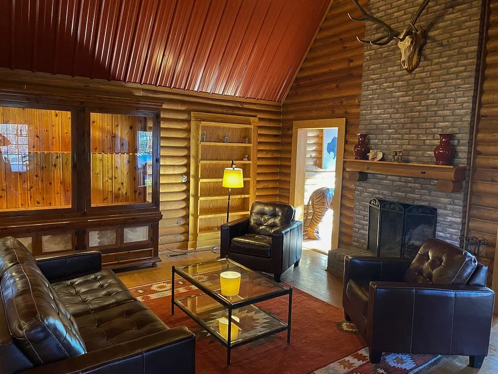 The interior of a cozy log cabin living room with leather furniture, a brick fireplace, wooden walls, and an animal skull with antlers mounted above the fireplace.
