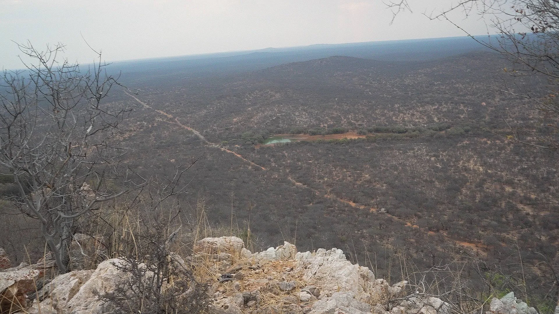 A vast, rocky, and barren landscape with sparse, leafless trees in the foreground. The terrain extends into the distance with a small body of water and a faint trail or dirt road visible across the land. The sky is overcast or hazy.
