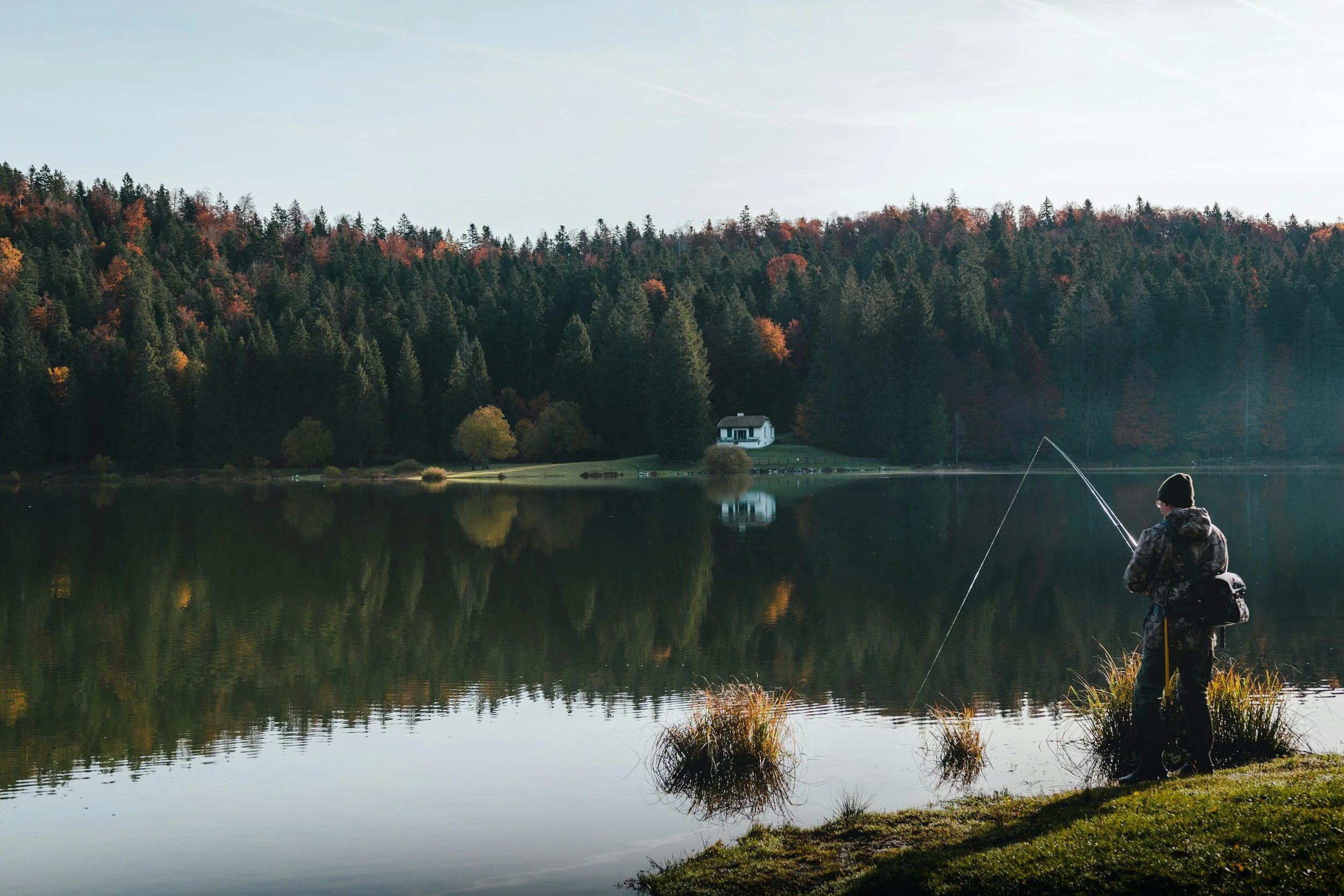 A person fishing by a lake with a forested hillside and a house in the background during autumn.