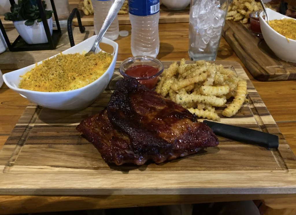 A meal consisting of ribs, seasoned French fries, mac and cheese with breadcrumbs, ketchup, and two glasses of water on a wooden table.