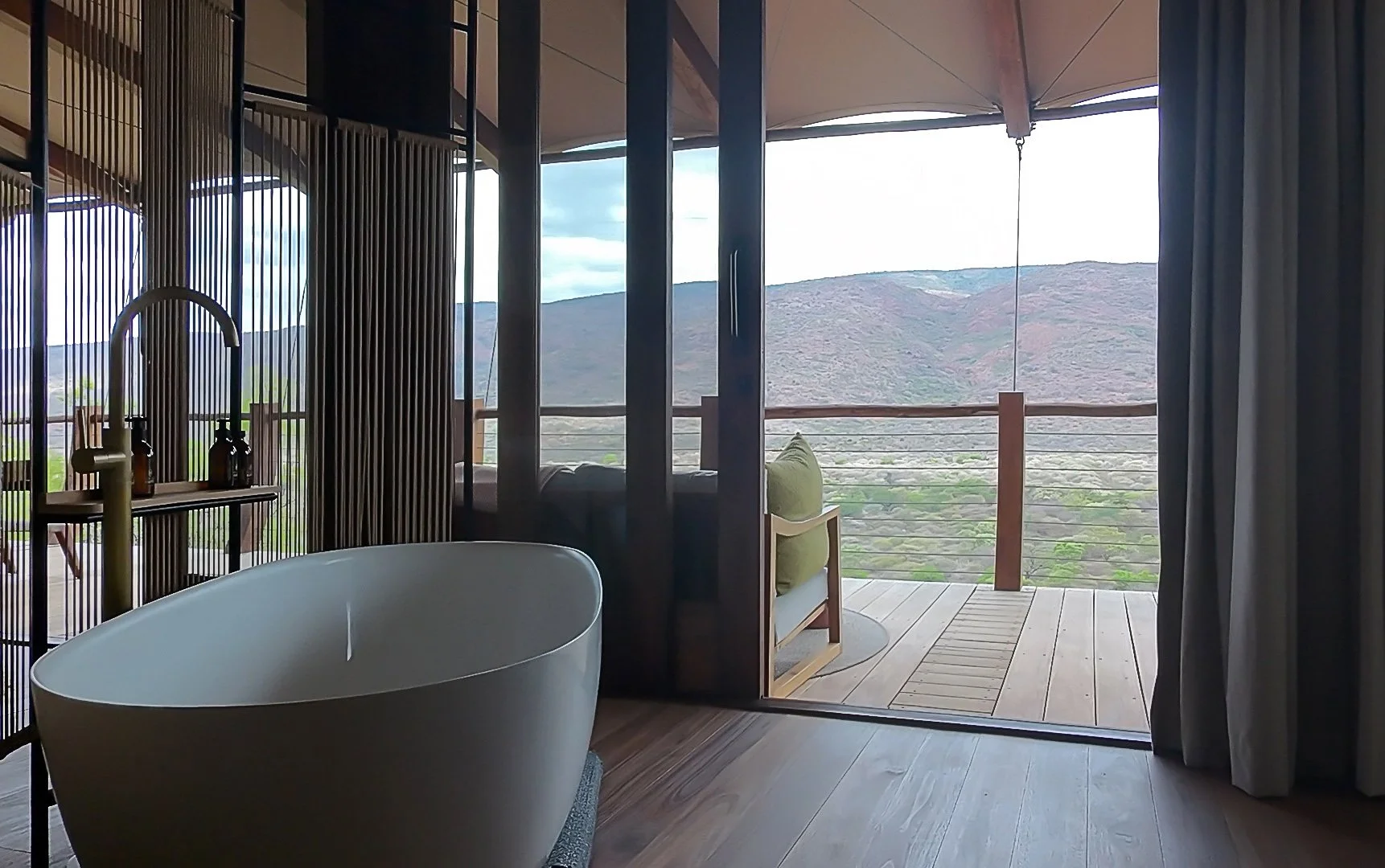 Freestanding bathtub inside a luxury safari suite with floor-to-ceiling windows opening to a wooden deck and panoramic views of a green valley and distant mountains.
