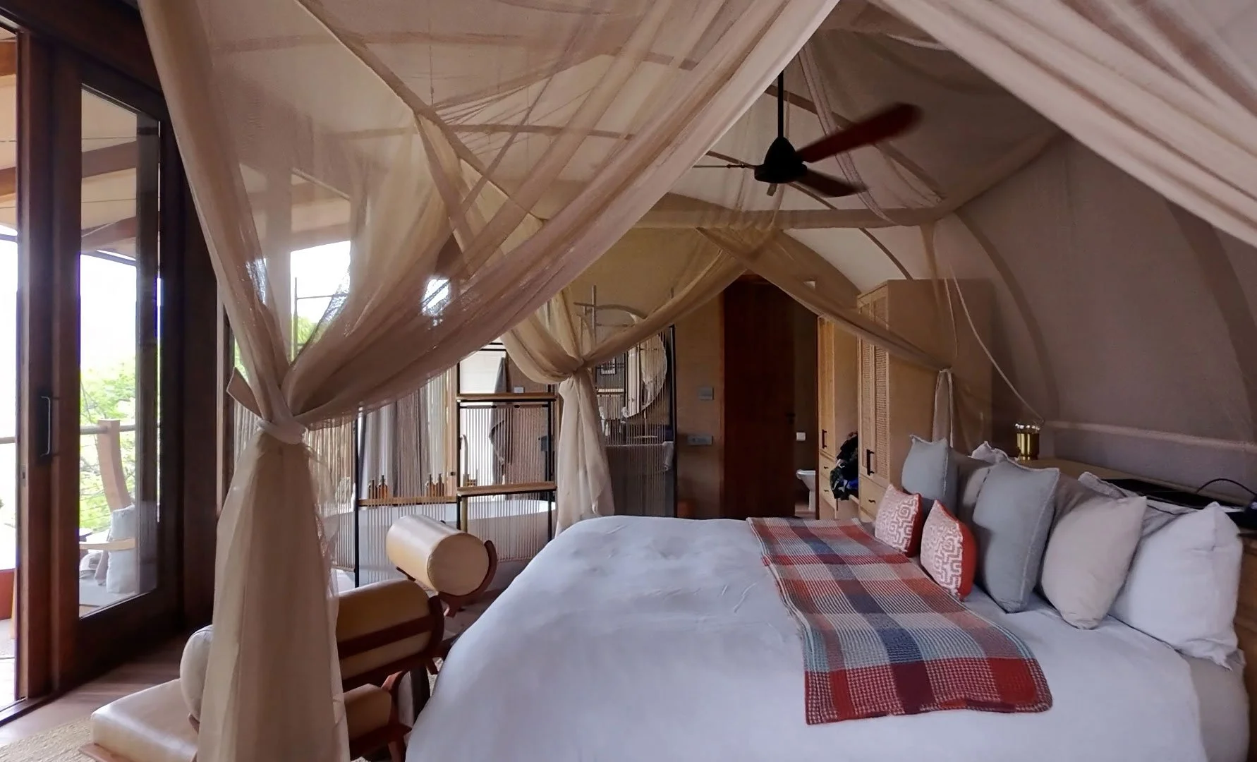 Luxury safari-style bedroom with a large canopy-draped bed, soft neutral décor, plush pillows, and natural light coming through glass doors leading to an outdoor deck.