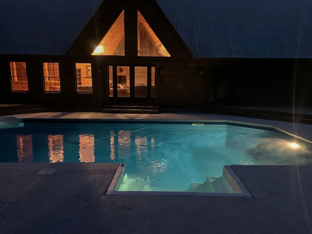 A nighttime view of a swimming pool in front of a wooden house with large windows and a steeply pitched roof, illuminated from inside.