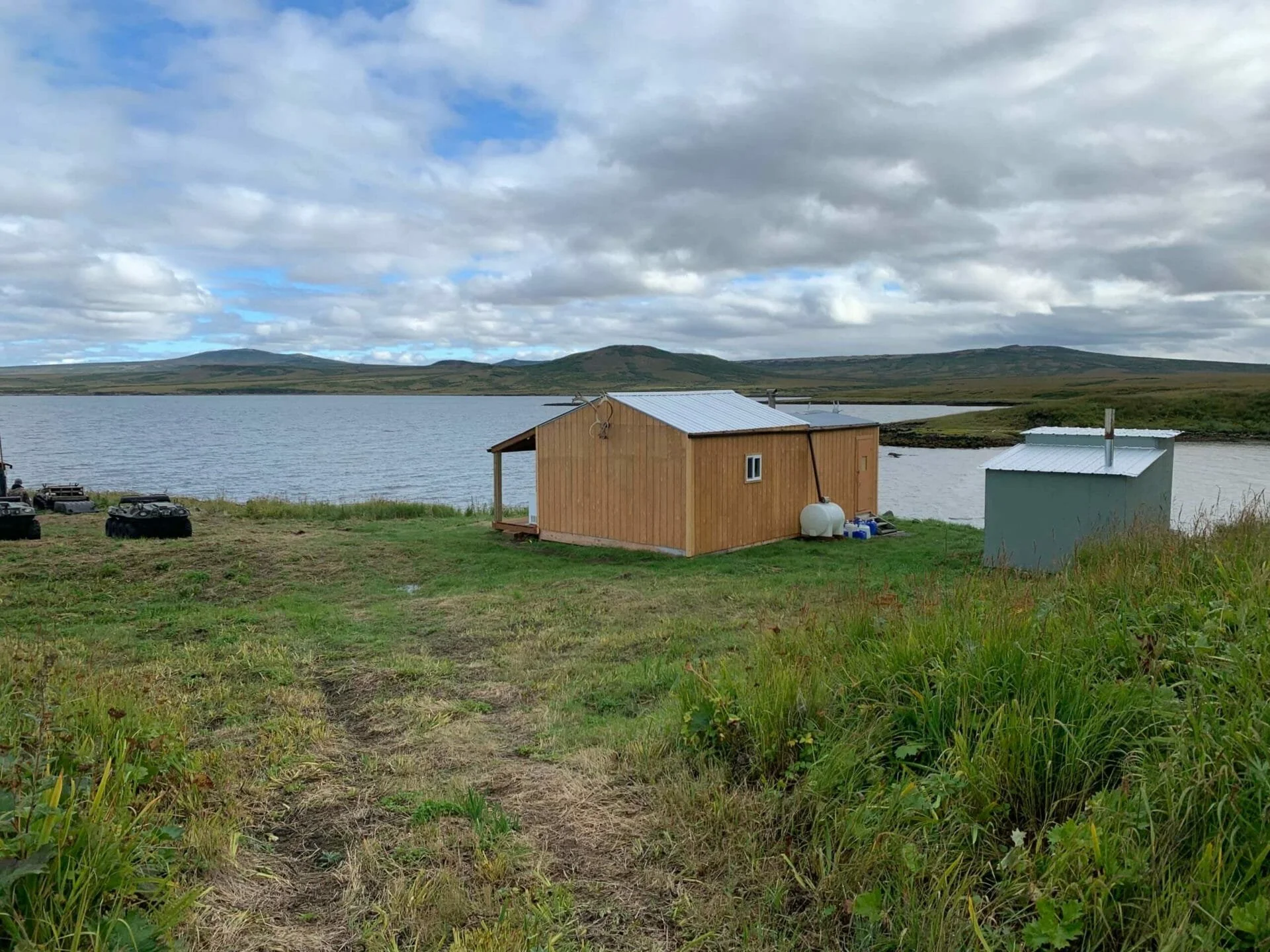 A small wooden cabin near a body of water with hills in the background, cloudy sky, and some parked vehicles nearby.